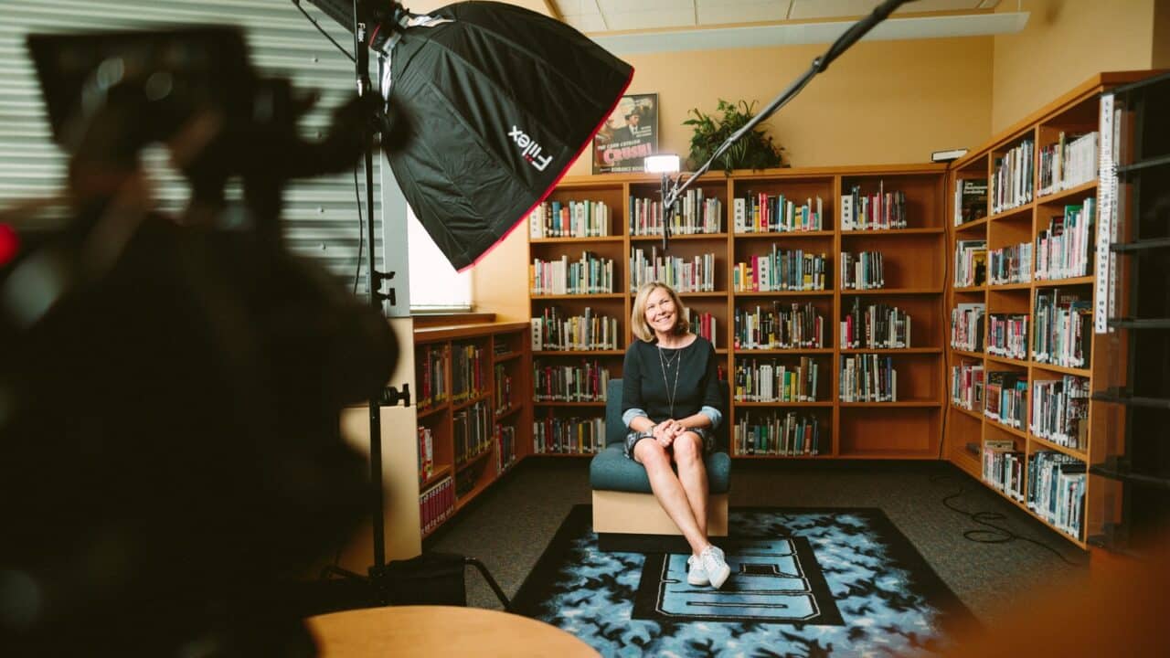 vrouw zittend op armloze stoel met licht tussen boekenkasten in kamer die een Skillshare cursus filmt.