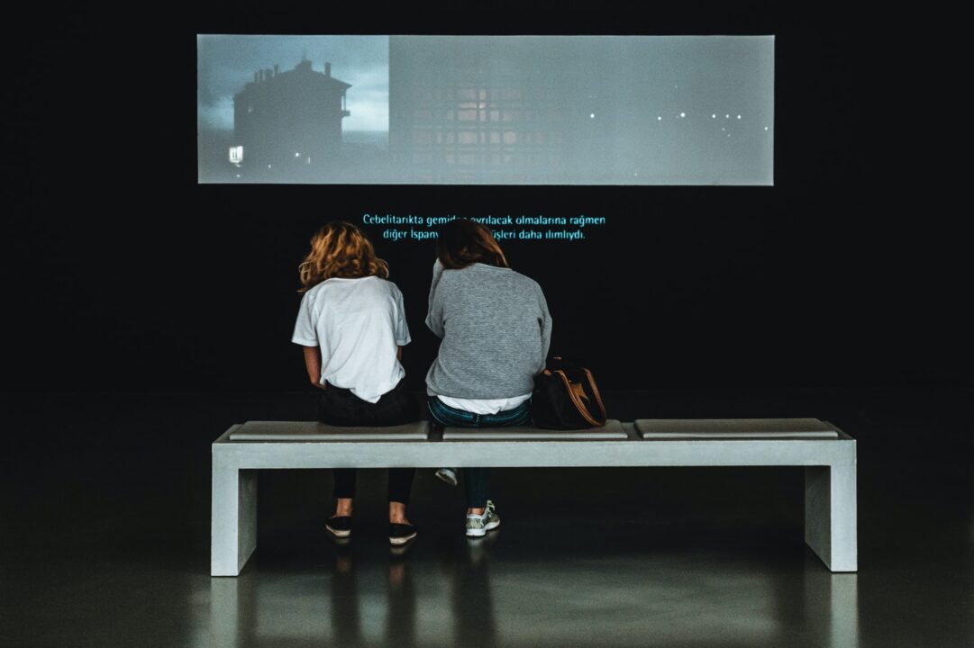 two women sitting on table watching a video with subtitles.