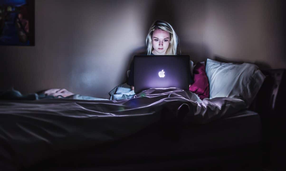 vrouw zittend op bed met MacBook op schoot. Ze is bezig met een Moodle Video.